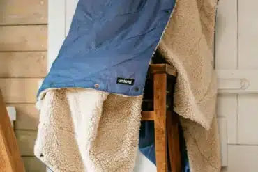 Blaue UNFOLD Kuscheldecke mit weicher Sherpa-Fleece-Seite über einem Holzstuhl
