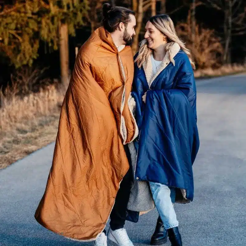 Zwei Personen tragen eine blaue und eine rostfarbene UNFOLD Kuscheldecke als Cape draußen auf einem Weg.