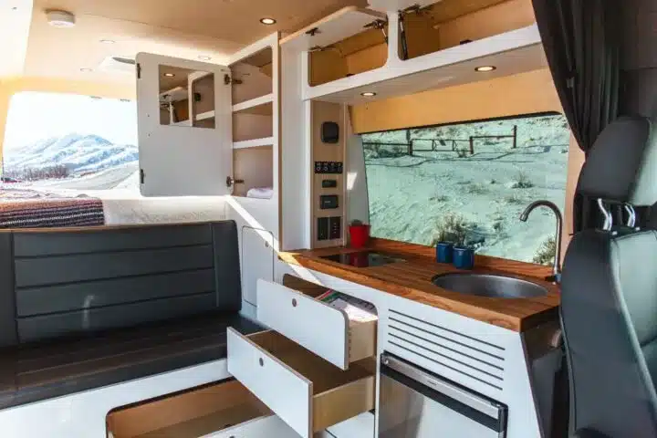 Innenraum eines Campervans mit Holzarbeitsplatte, Spüle, Schubladen, Sitzbank und Schlafbereich