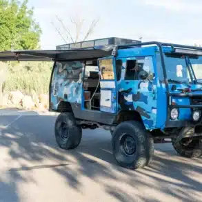 Blauer Volvo C303 TGB 1111 mit Camouflage-Folierung, groben Offroad-Reifen, Dachkorb und ausgefahrener Markise