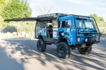 Blauer Volvo C303 TGB 1111 mit Camouflage-Folierung, groben Offroad-Reifen, Dachkorb und ausgefahrener Markise