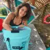 Türkiser YETI Hopper M20 Kühlrucksack mit geöffneter Kühlöffnung neben einer Frau im Garten