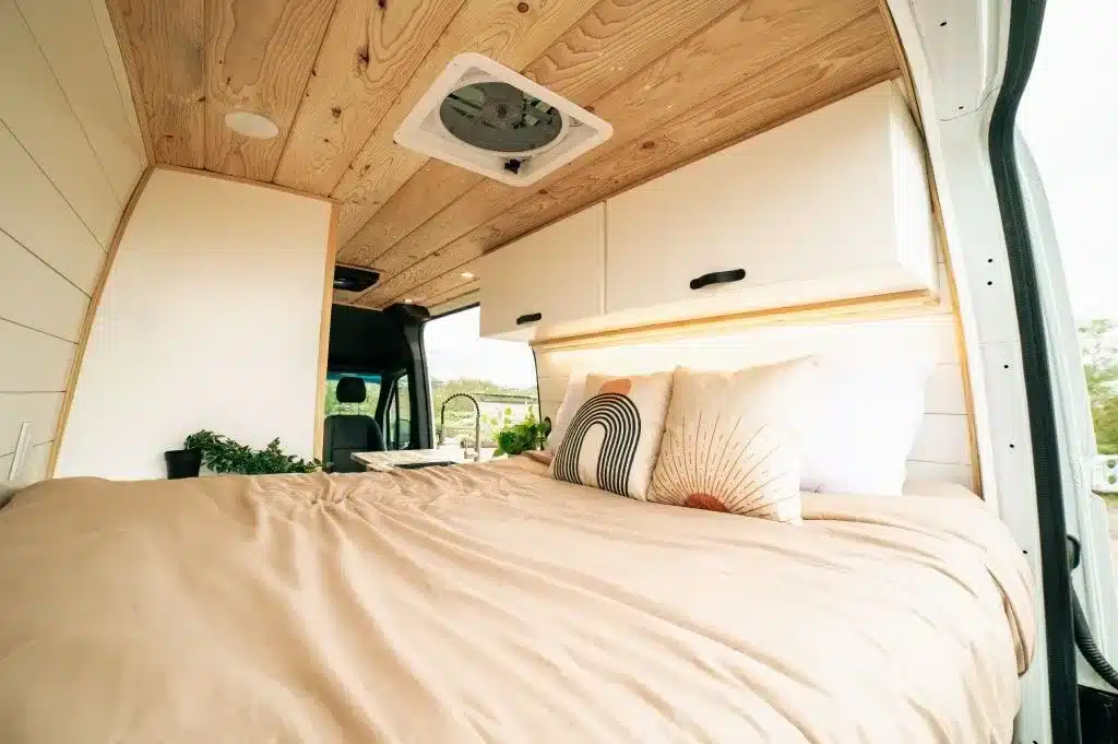 Innenraum des Papago Vans Zion mit breitem Bett, Holzhimmel, Oberschränken und Dachventilator