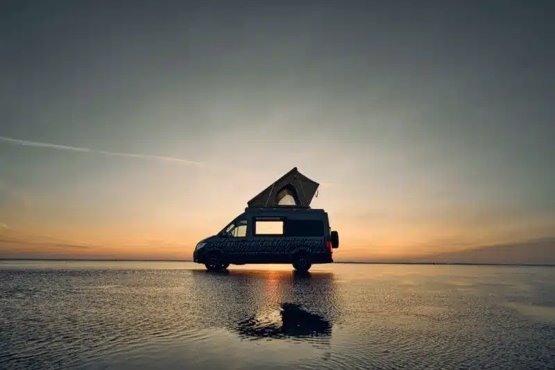 Dunkler Custom-Bus Camper XL mit geöffnetem Aufstelldach steht bei Sonnenuntergang im flachen Wasser