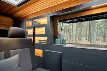 Innenraum des Custom-Bus Camper XL mit dunkler Küchenzeile, Holzdecke und großem Fenster mit Waldblick