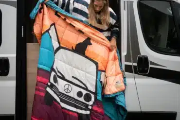 Frau hält eine farbenfrohe All-Terrain-Bettdecke an der geöffneten Schiebetür eines weißen Vans.